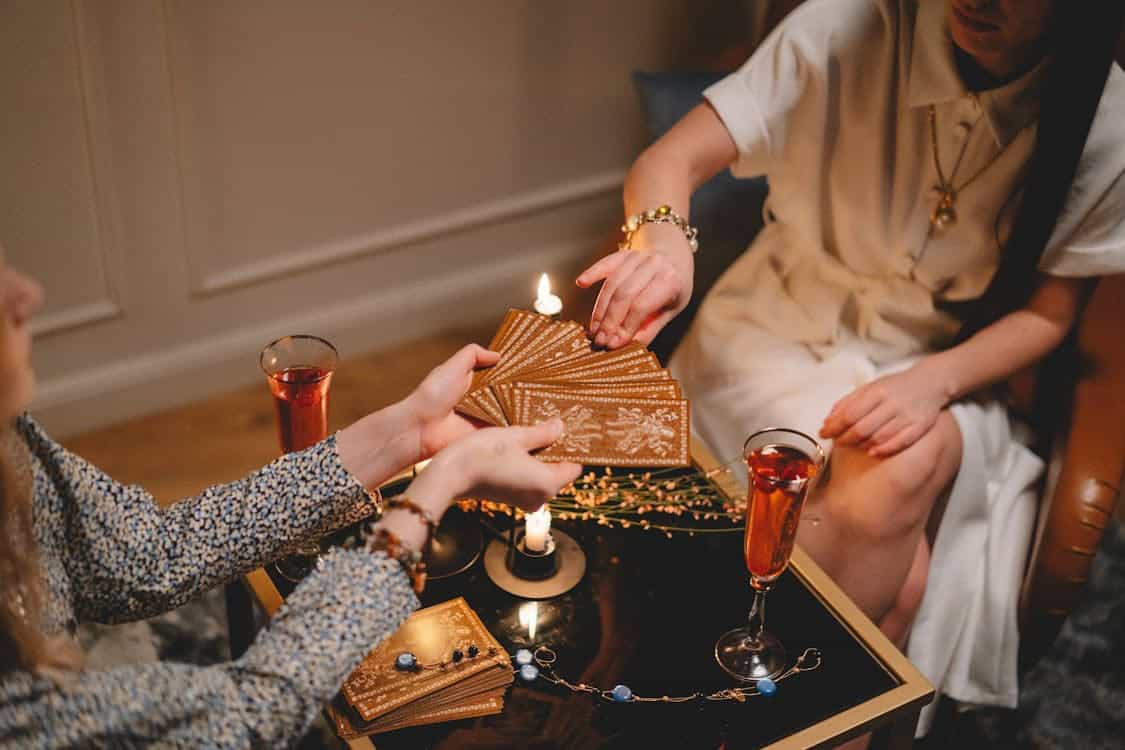 Two women sit at a table with tarot cards, lit candles, and drinks. One woman fans out the cards while the other chooses one, suggesting a fortune-telling or tarot reading session.