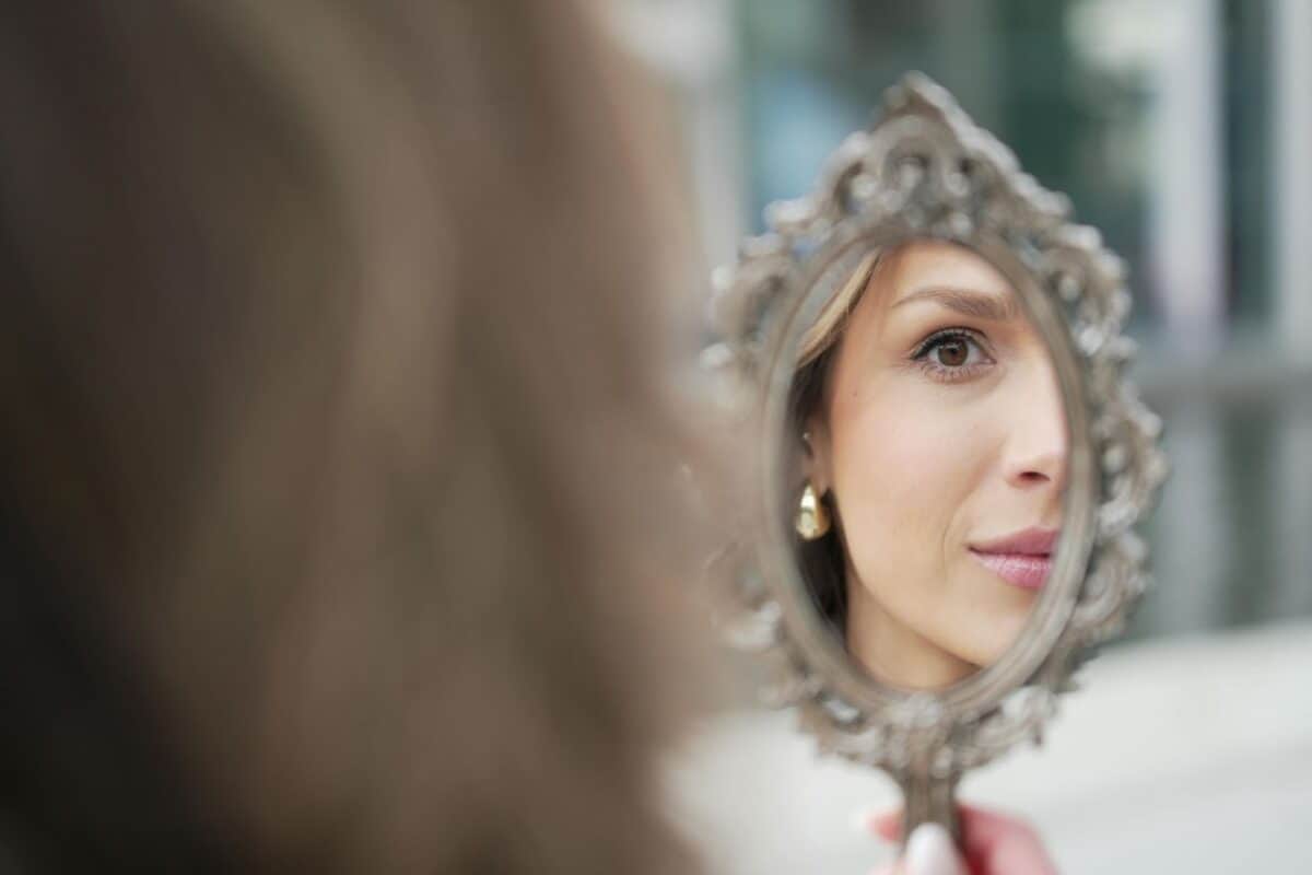 A woman holds an ornate hand mirror, reflecting her face thinking about wellness. Only part of her face is visible in the mirror, with a focus on her eye and a gentle smile. The background is softly blurred.