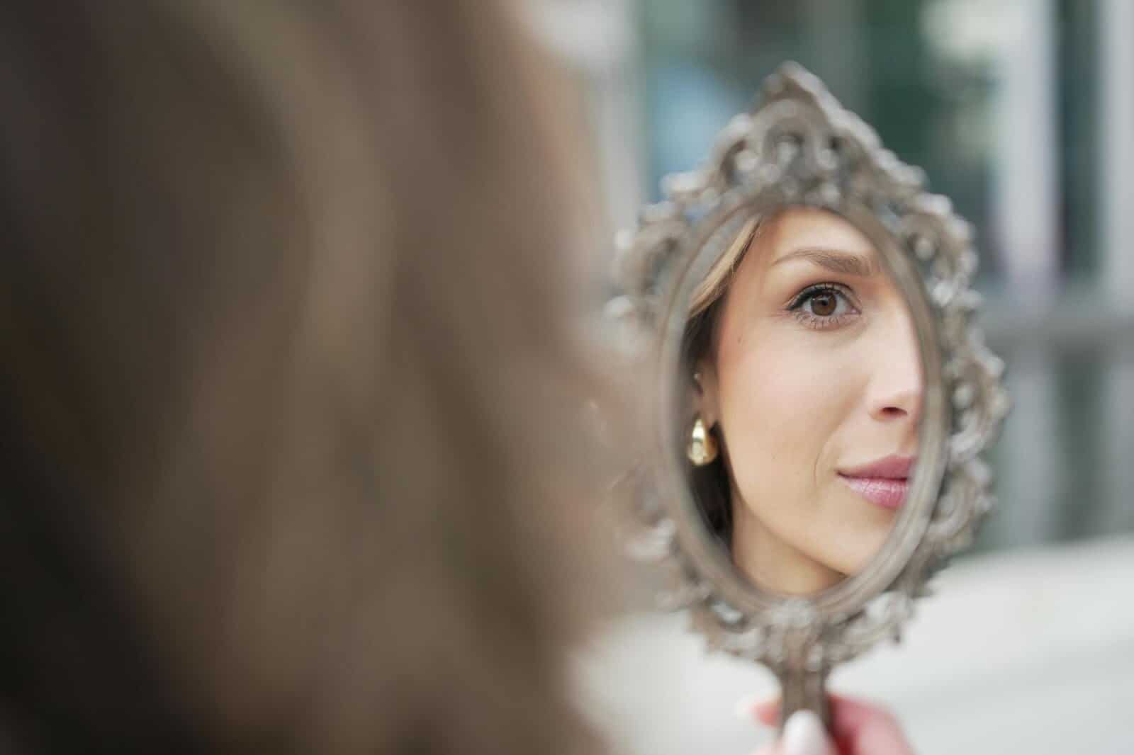A woman holds an ornate hand mirror, reflecting her face. Only part of her face is visible in the mirror, with a focus on her eye and a gentle smile. The background is softly blurred.