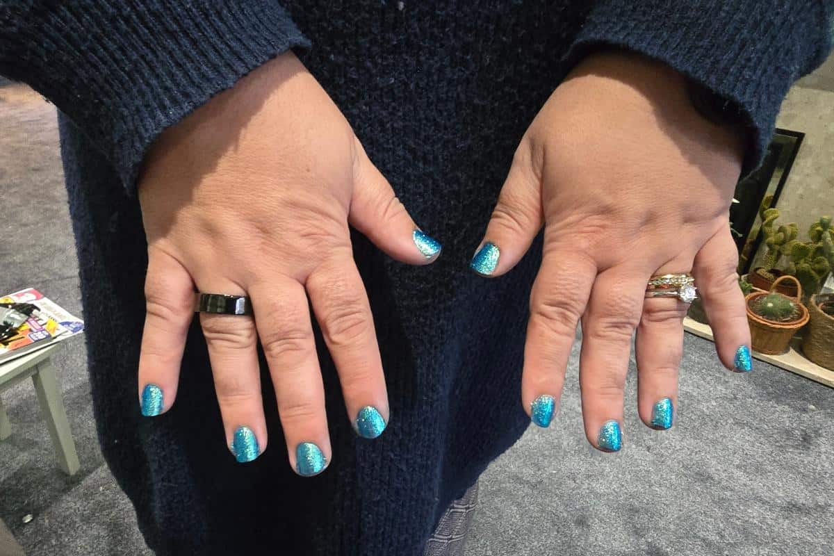 A person wearing a dark blue sweater displays their hands with short, blue glittery manicured nails and several rings, including the Ultrahuman Ring Air, on both hands. The background features a gray carpet, plants, and a small white table.