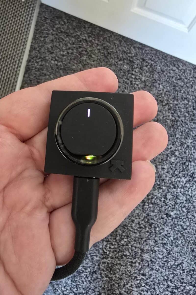 A hand holding a small black electronic device with a circular dial, an illuminated green light, and a connected black cable—possibly part of an Ultrahuman Ring Air smart ring review. The background shows gray carpet and part of a white door.