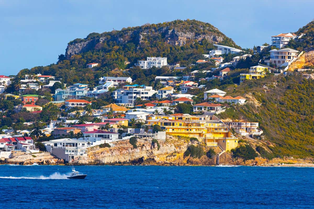 Colorful houses dot a hillside overlooking the blue sea on the French side St. Martin, with a speedboat gliding across the water and lush green hills providing a stunning backdrop.