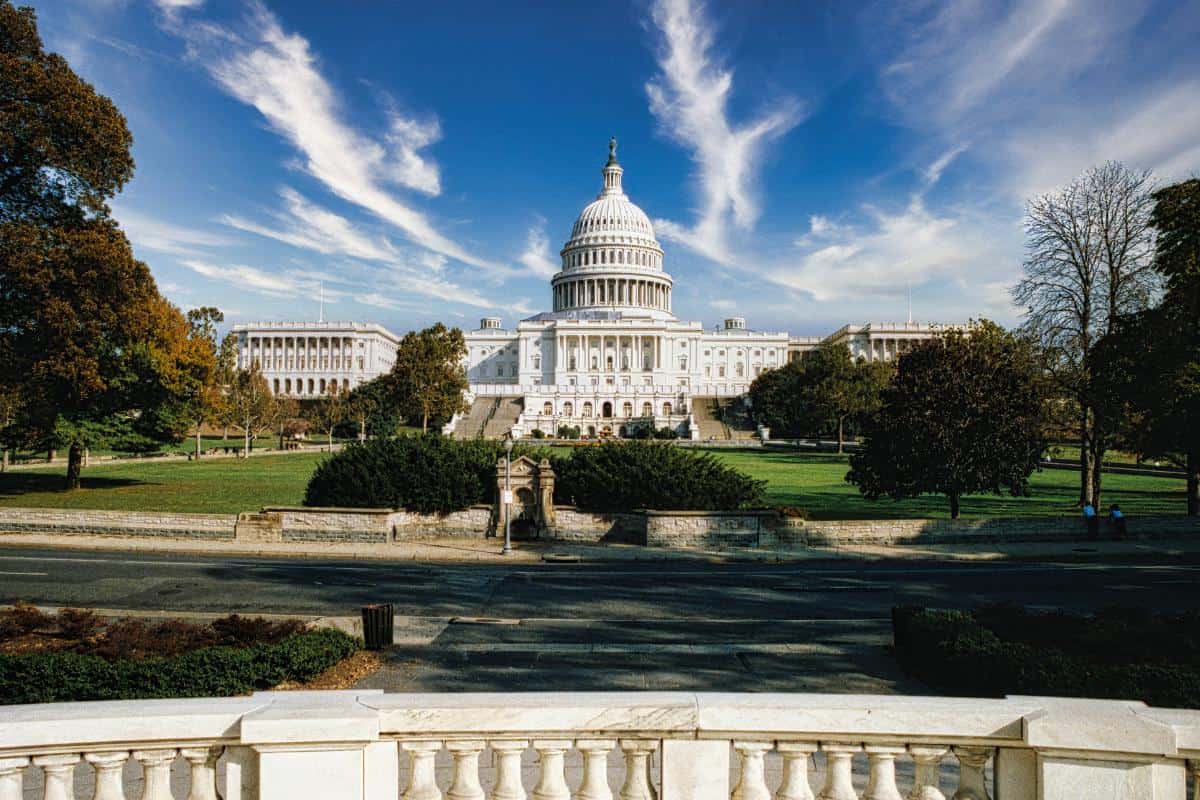 The U.S. Capitol building, one of Washington DC’s iconic landmarks, stands across a green lawn with trees and a stone railing in the foreground, beneath a blue sky with wispy clouds.