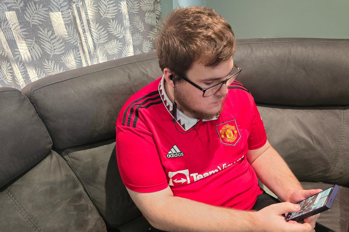A man wearing glasses and a red Manchester United jersey sits on a gray couch, looking down and using his smartphone—perhaps browsing for the latest tech gifts. Light filters in through patterned curtains behind him.