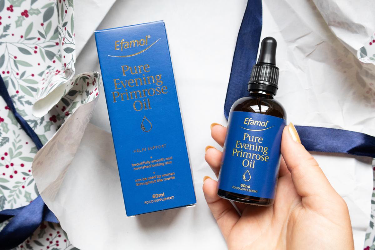 A hand holds a bottle of Efamol Pure Evening Primrose Oil with a dropper, next to its blue and gold packaging, placed on crumpled white paper with a blue ribbon—perfect as a Secret Santa or stocking filler—floral-patterned paper in the background.