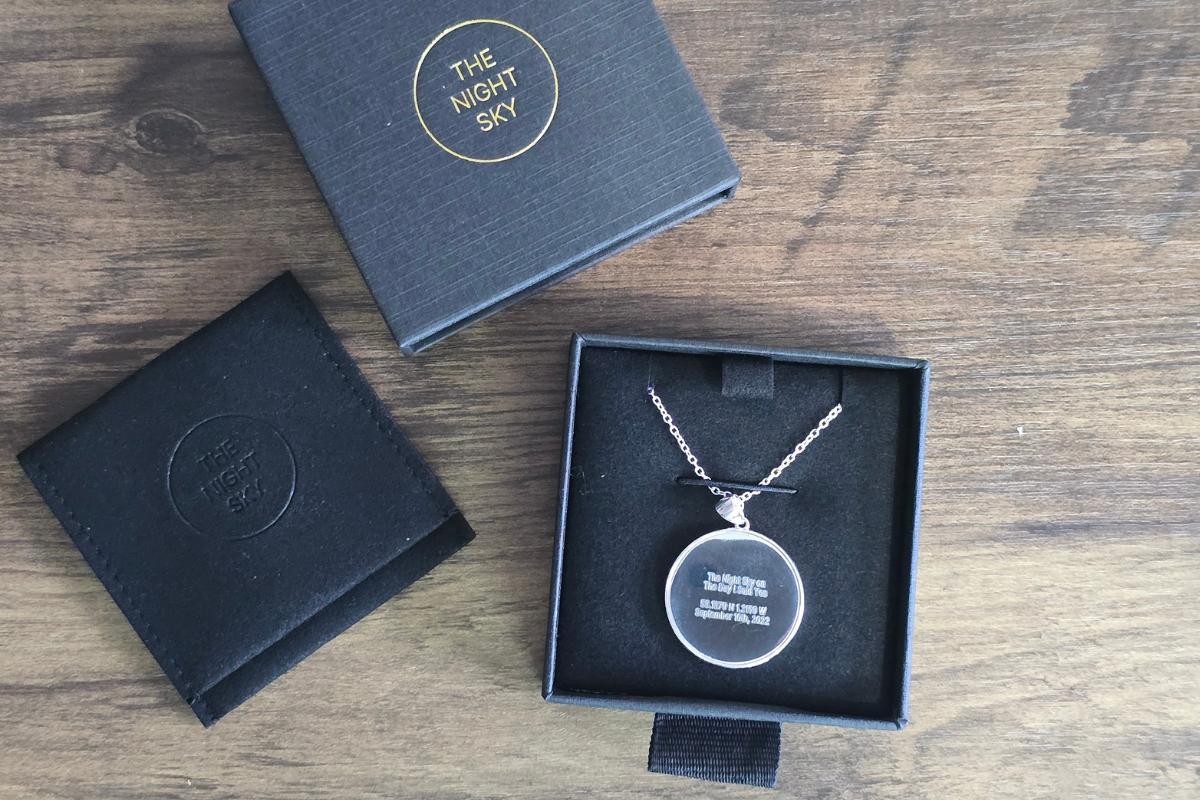 A silver night sky necklace with a round pendant in a black box, next to another closed black box, both labeled "The Night Sky," displayed on a wooden surface—perfect for holiday gifts and featured in our Christmas Gift Guide.