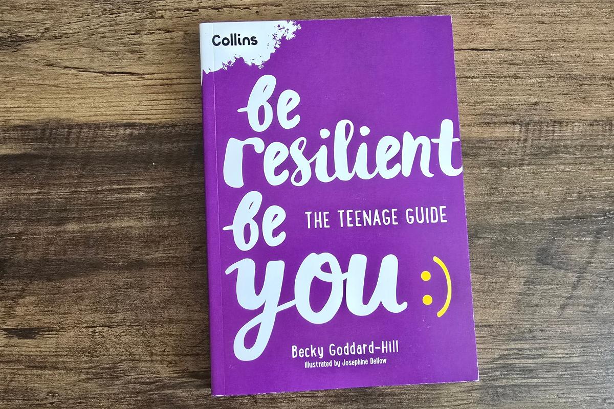 A purple book titled “Be Resilient: The Teenage Guide” by Becky Goddard-Hill lies on a wooden surface—an inspiring pick for any children's Christmas gifts or Christmas gift guide. The cover features white and yellow text, a smiley face, and the Collins logo.