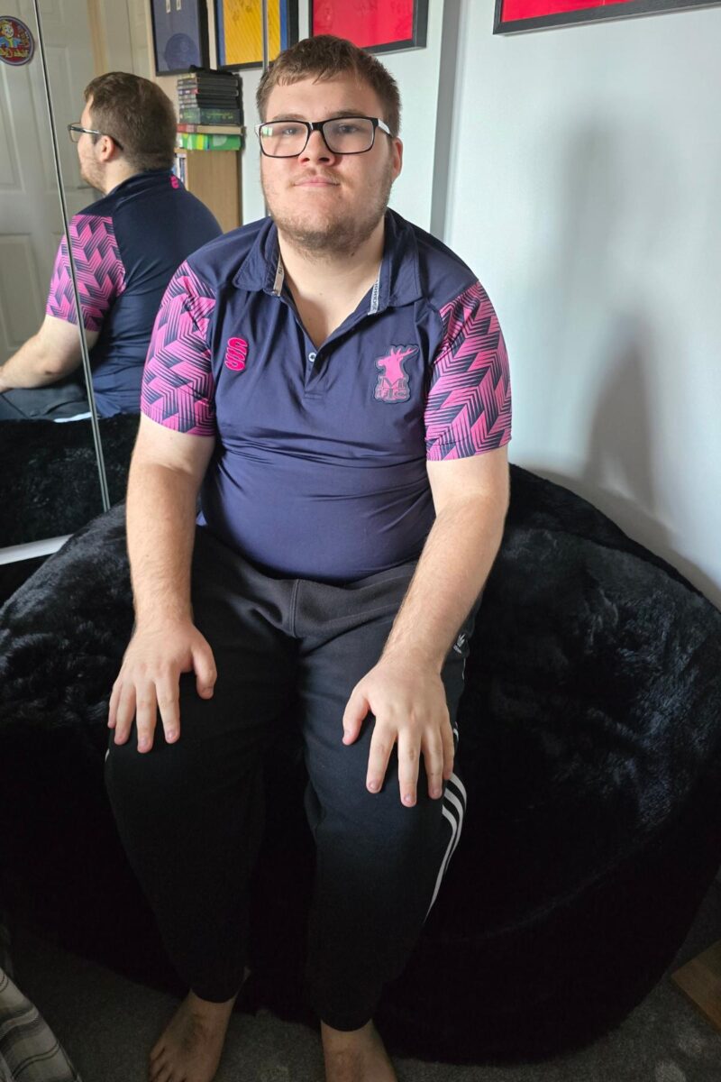 A young man with short hair, glasses, and a beard sits barefoot on a black Monster Bean Bag chair, wearing a navy and pink sports shirt and black pants. His reflection is visible in the mirror behind him—a real-life bean bag review scene.