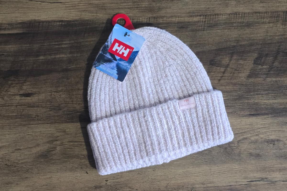 A light beige knitted beanie with a folded brim and a Helly Hansen tag lies on a wooden surface—perfect for any Christmas Gift Guide or thoughtful Christmas gifts.