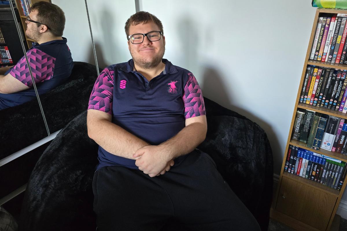 A smiling man with glasses sits on a Monster Bean Bag, wearing a navy and pink sports shirt. A mirrored wardrobe and a bookshelf filled with DVDs and books are visible in the background.