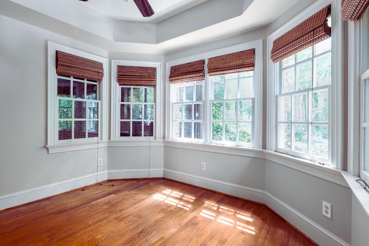 A bright, empty room with hardwood floors, light gray walls, and large bay windows featuring woven wood blinds that let in natural sunlight. Trees are visible outside, creating a connection to healthier spaces and harmonious indoor living.
