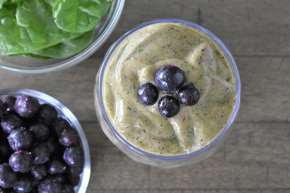 A glass of spinach smoothie topped with blueberries sits next to bowls of fresh spinach leaves and frozen blueberries on a wooden surface.
