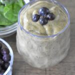A creamy spinach smoothie in a clear glass, topped with four blueberries. Nearby are bowls of fresh spinach leaves and blueberries, all arranged on a wooden surface.