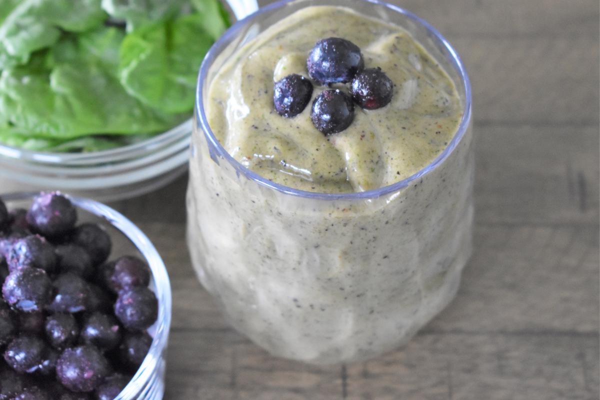 A creamy spinach smoothie in a clear glass, topped with four blueberries. Nearby are bowls of fresh spinach leaves and blueberries, all arranged on a wooden surface.
