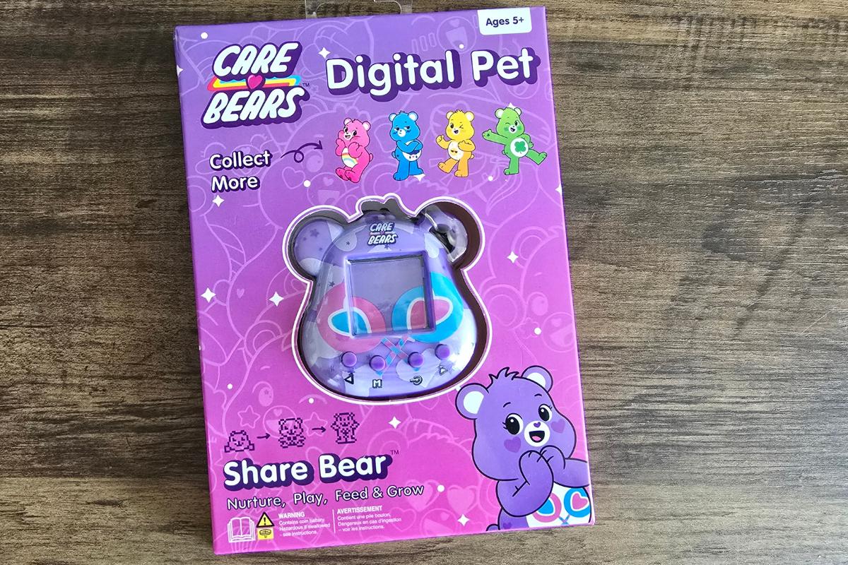 A boxed Care Bears Digital Pet toy featuring Share Bear, with colorful illustrations on a purple background. Perfect as a Stocking Filler or Secret Santa gift, this toy says “Nurture, Play, Feed & Grow” and is for ages 5+. The toy is visible in the box.