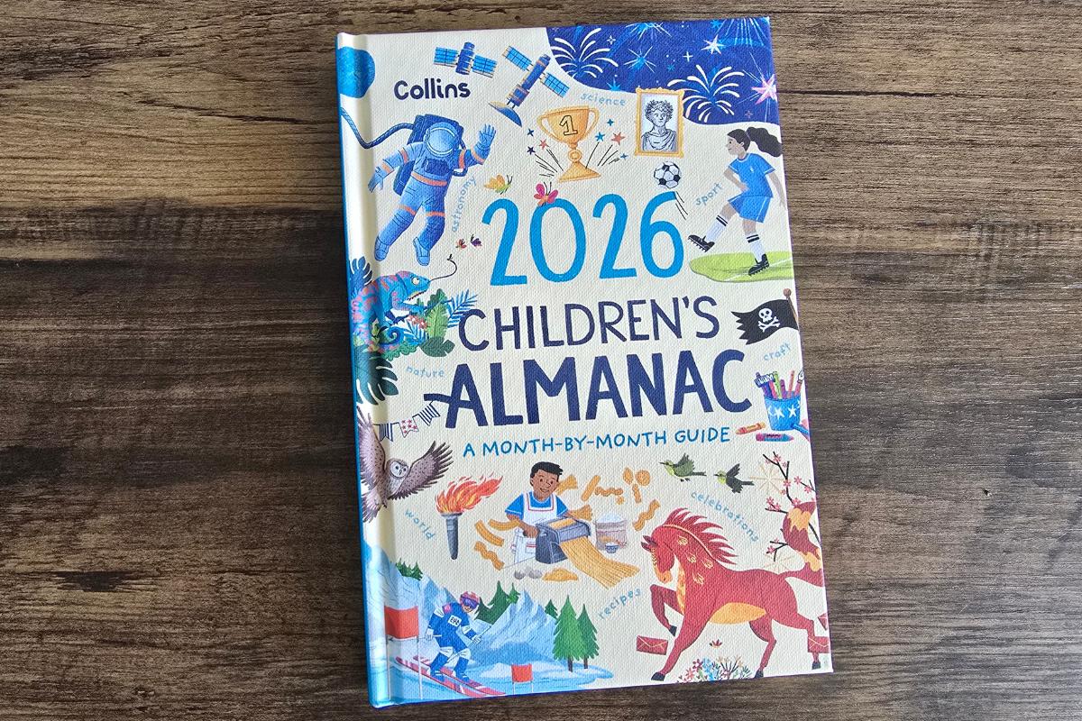 A colorful book titled "2026 Children's Almanac: A Month-by-Month Guide" by Collins lies on a wooden surface—an ideal pick for any children's Christmas gifts guide, with playful cover art of kids, animals, sports, an astronaut, and fireworks.