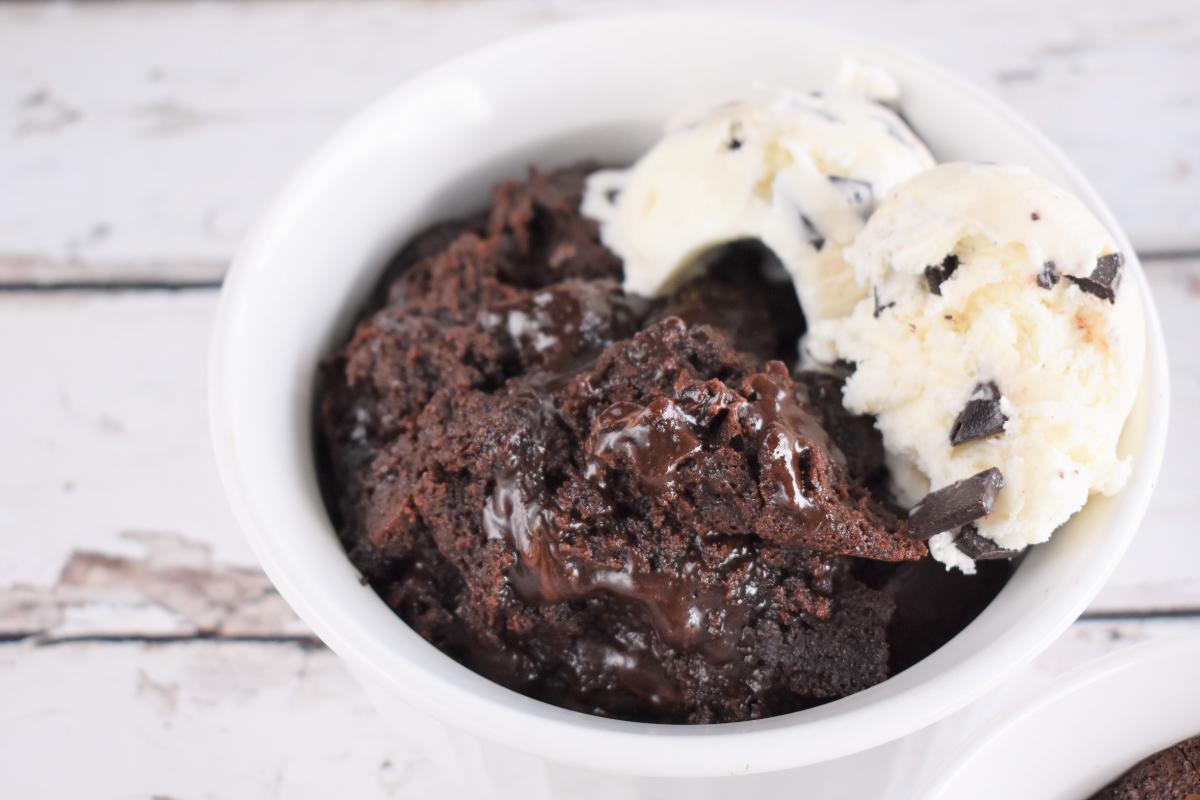A white bowl filled with gooey slow cooker chocolate cake, topped with two scoops of vanilla ice cream with chocolate chunks, set on a rustic white wooden surface.