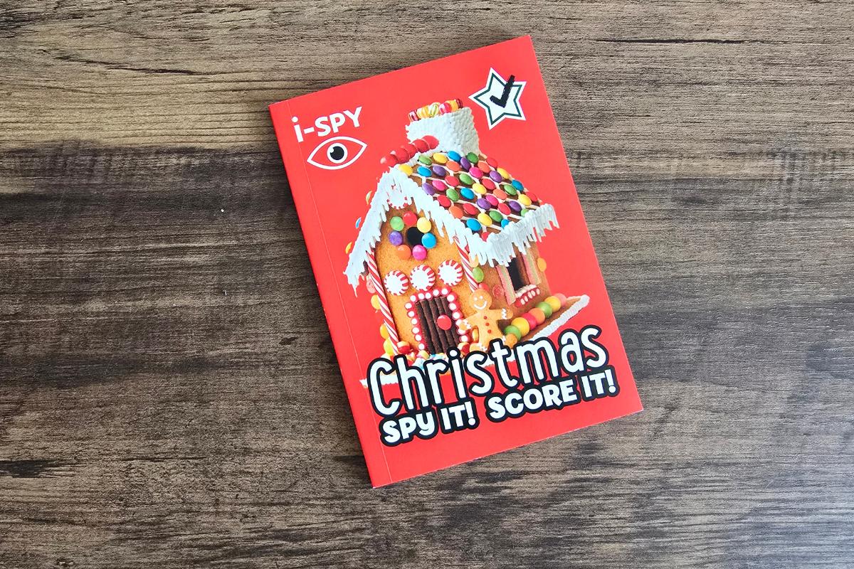 A red book titled "i-SPY Christmas Spy It! Score It!" featuring a decorated gingerbread house with candy, resting on a wooden surface—perfect for children’s Christmas gifts or adding to your Christmas gift guide.