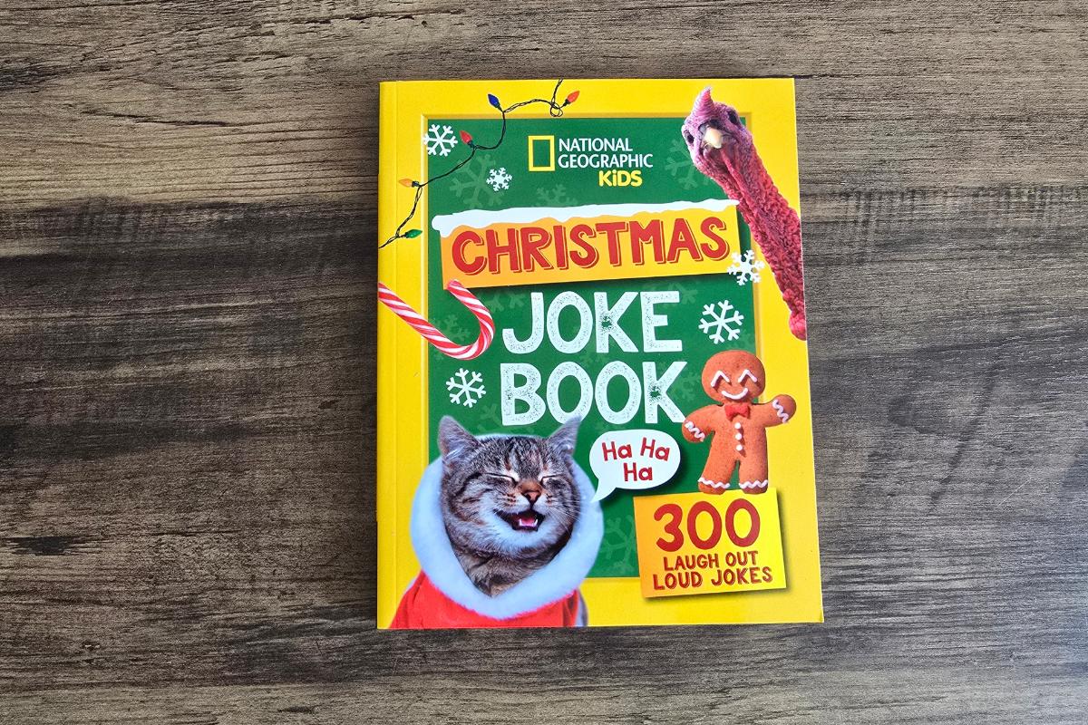 A children's book titled "National Geographic Kids Christmas Joke Book" lies on a wooden surface. Featured in the children's Christmas gift guide, its festive cover boasts a laughing cat in a Santa suit, a gingerbread man, and 300 laugh-out-loud jokes.