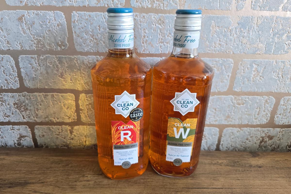 Two bottles of Clean Co non-alcoholic spirits, labeled "R" and "W," stand on a wooden surface against a light brick wall—perfect additions to your Christmas Gift Guide for Him. Both amber bottles feature blue caps and white labels.