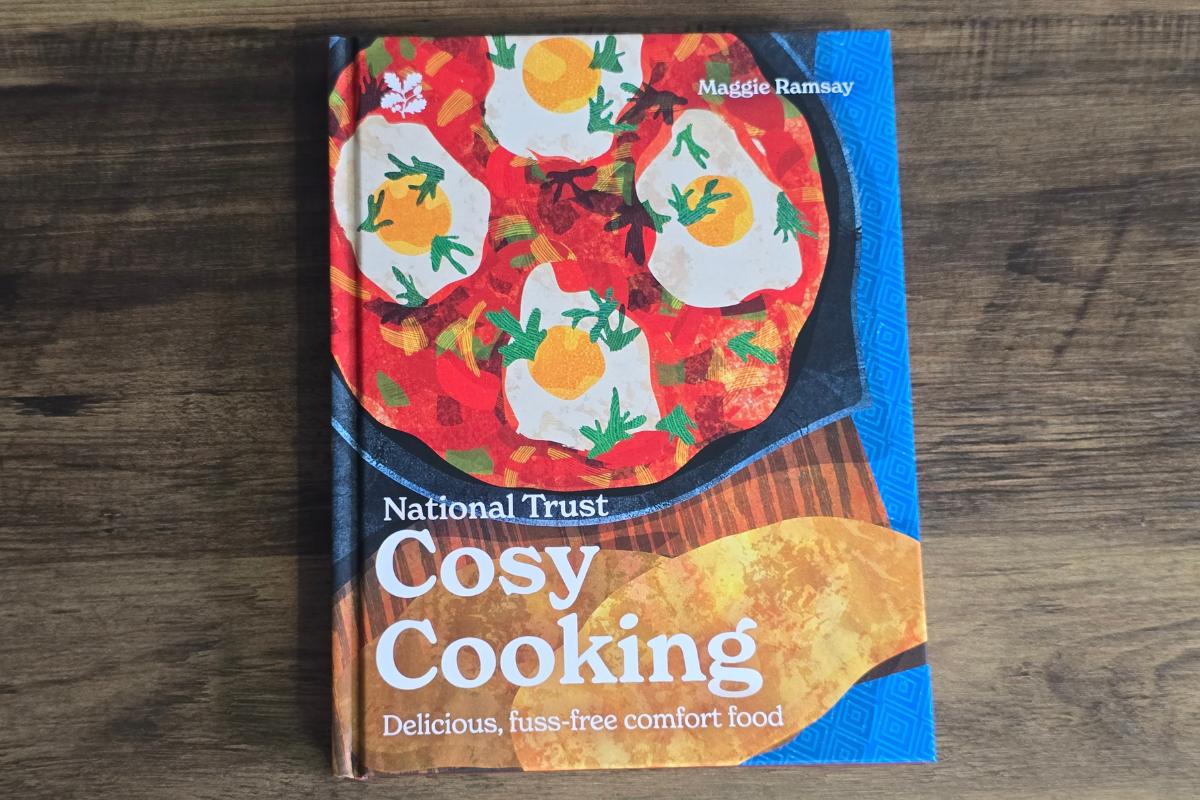 A cookbook titled "Cosy Cooking: Delicious, fuss-free comfort food" by Maggie Ramsay is placed on a wooden surface. The cover features an illustration of a skillet with eggs, tomato sauce, and herbs—perfect for Christmas Presents.