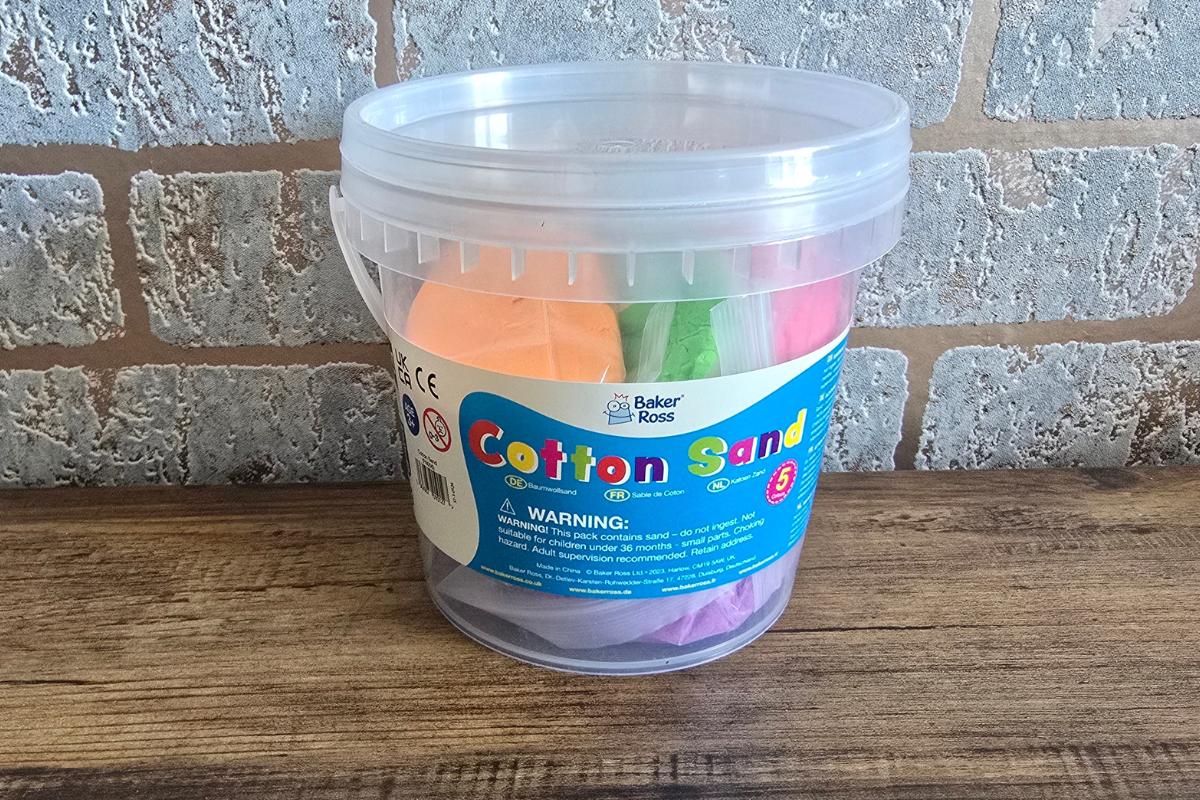 A clear plastic tub labeled "Cotton Sand" by Baker Ross sits on a wooden surface, filled with colorful sand in orange, green, pink, and purple—perfect for any children's Christmas gift guide. The background is a brick wall.
