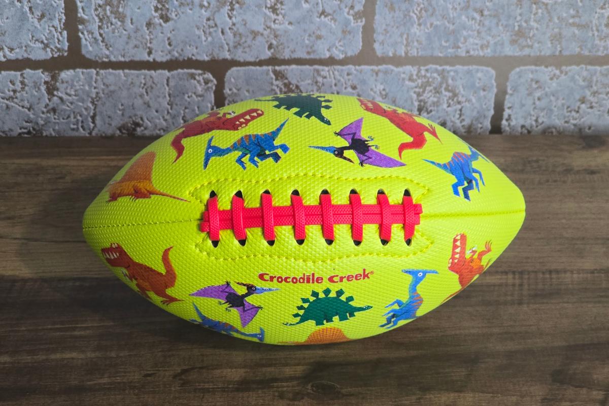 A yellow football with red laces, featuring colorful dinosaur illustrations and "Crocodile Creek," sits on a wooden surface with a brick wall backdrop—an ideal pick for any children's gift guide.