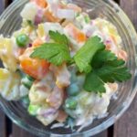 A bowl of Vegan Czech Potato Salad featuring peas, carrots, potatoes, and onions in creamy dressing, garnished with fresh parsley, on a wooden surface.