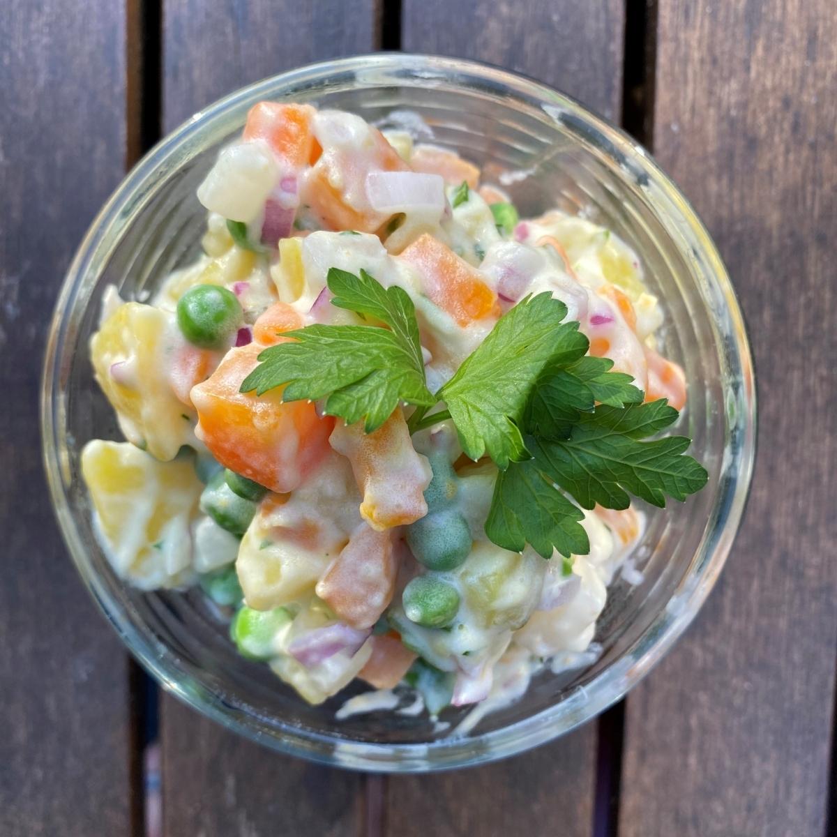 A glass bowl filled with creamy Vegan Czech Potato Salad, featuring diced carrots, potatoes, peas, and corn, garnished with fresh parsley. The bowl sits on a wooden surface.