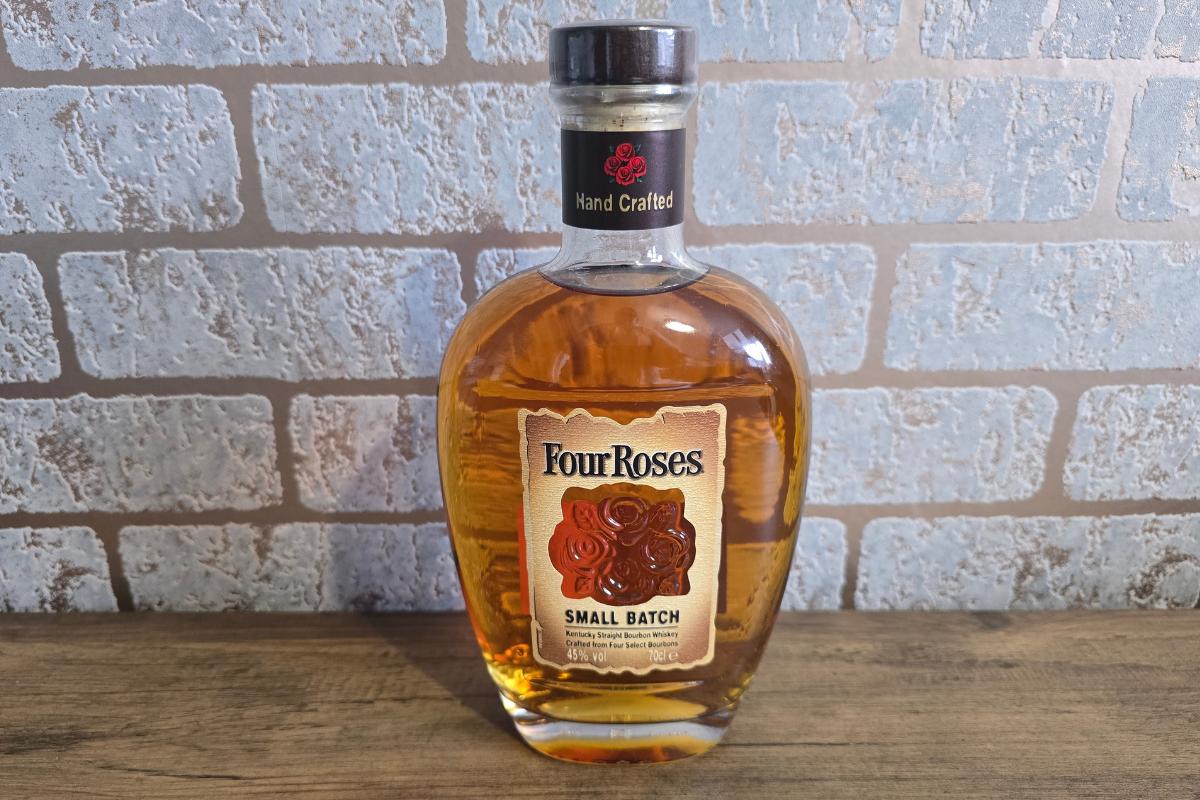 A bottle of Four Roses Small Batch bourbon stands on a wooden surface against a light brick wall—an elegant pick for any Christmas Gift Guide, with its clear label, red roses, and metallic cap.