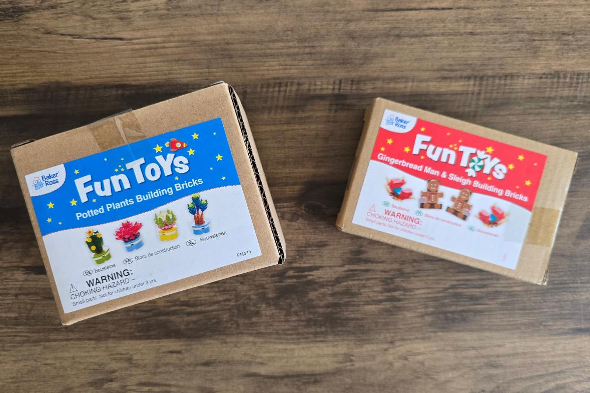 Two cardboard boxes labeled "Fun Toys" on a wooden surface; one holds potted plants building bricks, and the smaller box contains gingerbread men and sleigh bricks—perfect additions to any kids gift guide this holiday season.