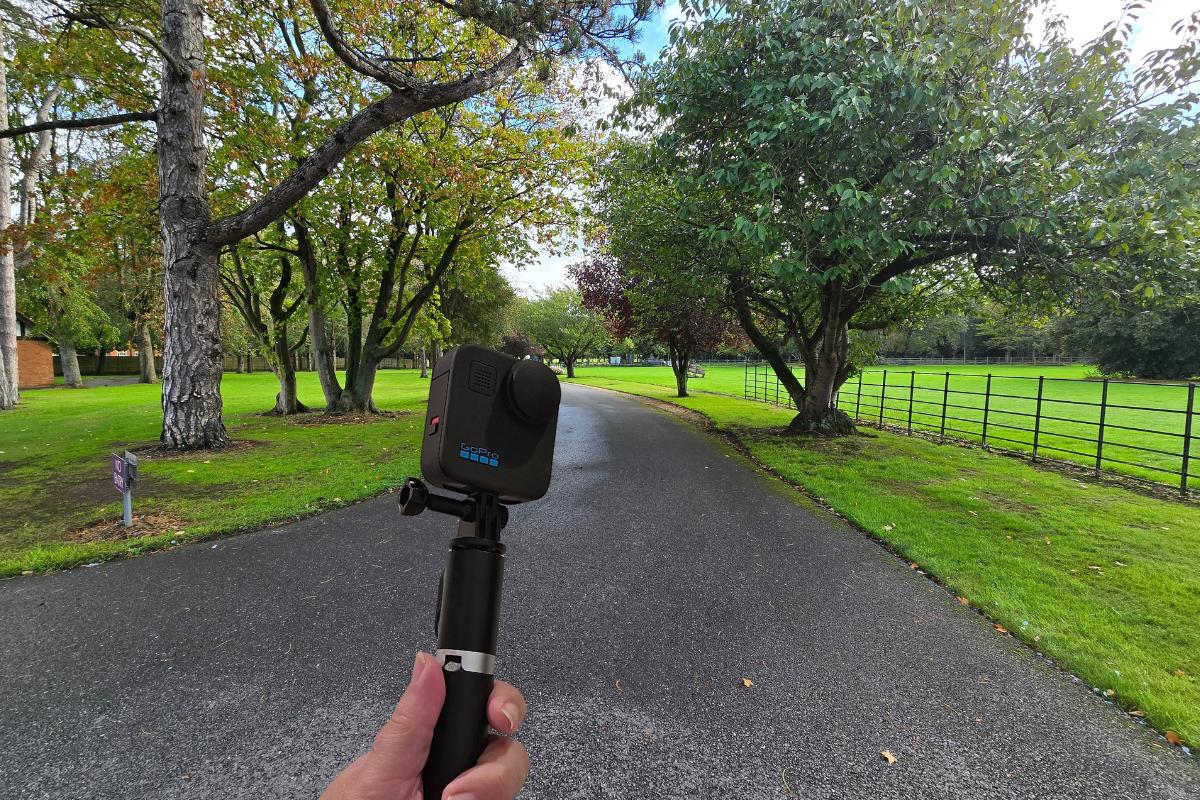 A hand holds a GoPro camera on a selfie stick, capturing a paved path lined with green grass and trees in a park on a bright day—perfect inspiration for tech gifts for outdoor adventurers.