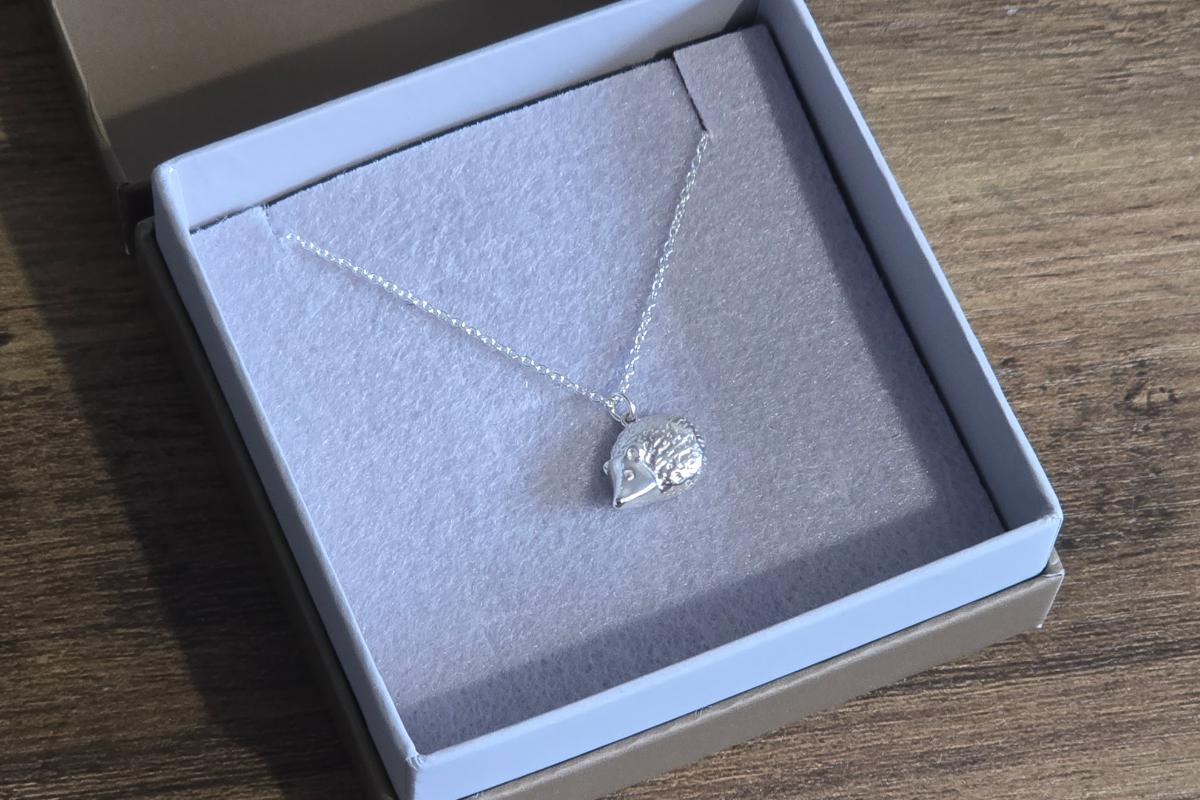 A silver necklace with a hedgehog-shaped pendant is displayed in an open gray jewelry box, making it a charming choice from the Christmas Gift Guide for Her, set on a wooden surface with soft, light gray inner lining.