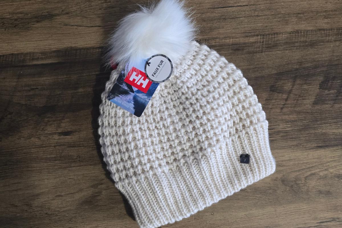 A cream-colored knit beanie with a fluffy white pom-pom, displayed on a wooden surface. With ribbed edging and festive tags, it's a cozy pick for the Christmas Gift Guide or as one of this season's top Christmas gifts for her.