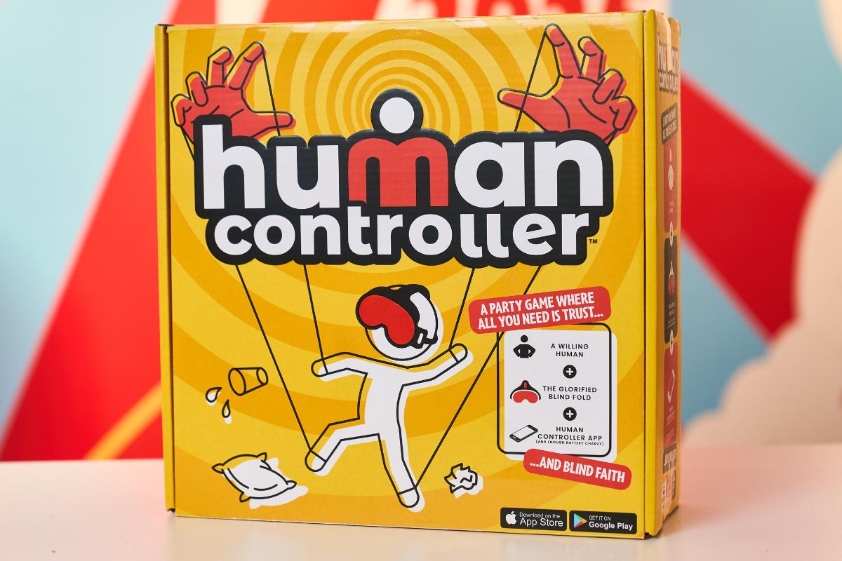 A yellow game box labeled "Human Controller" features a cartoon figure being controlled by puppet strings and large red hands above. Perfect for kids, this party game is sure to be on every Christmas List of Top Toys 2025, with fun depictions of game elements.
