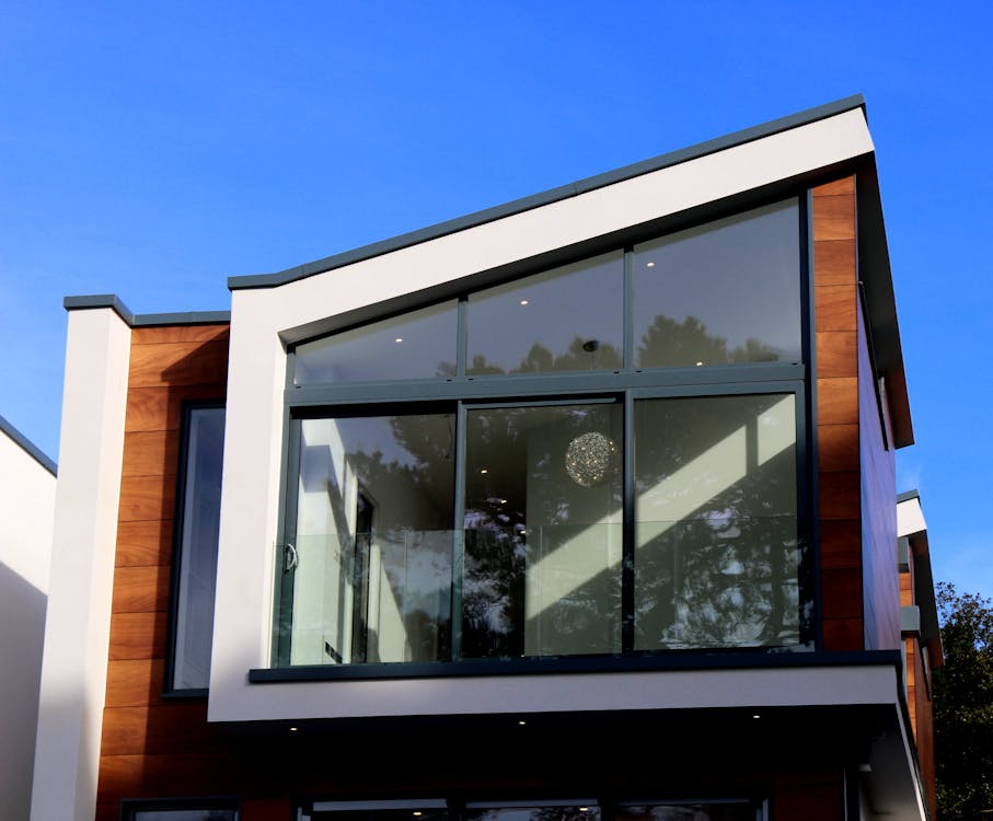 Modern two-story house exterior with large floor-to-ceiling windows, wood and white paneling, and a slanted roof against a clear blue sky. Tree reflections are visible in the glass.
