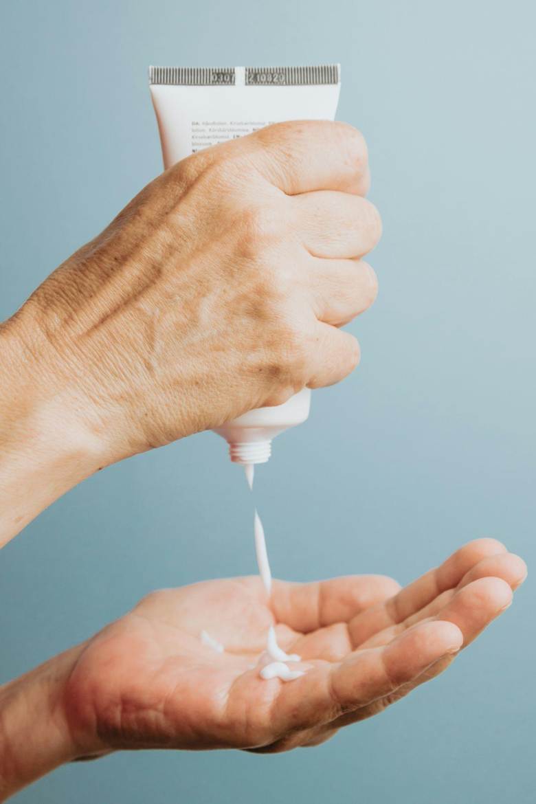 A person squeezes white lotion from a tube onto the palm of their hand against a plain light blue background.