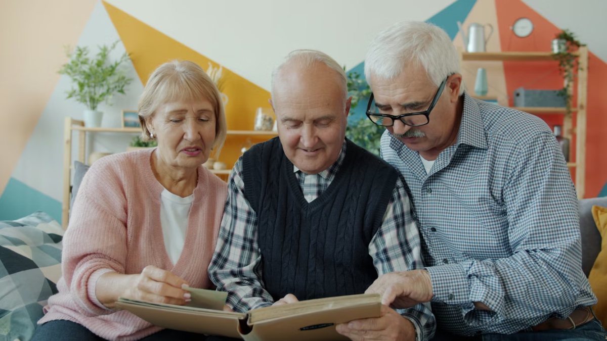 Three older adults sit closely together on a sofa, smiling and looking through a photo album in a cosy living room with shelves and plants in the background.