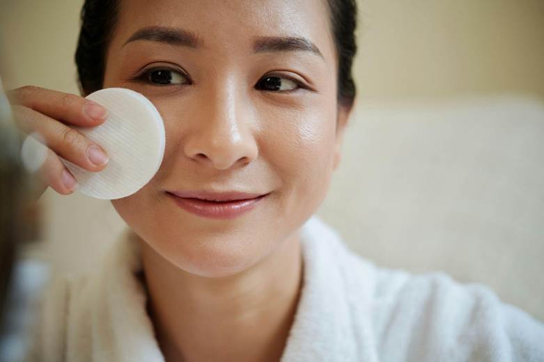 A woman in a white robe smiles while gently cleansing her face with a cotton pad.