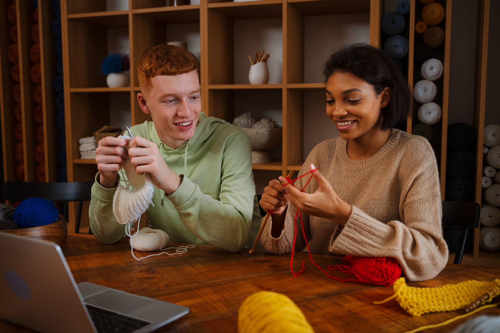 Two people sit at a wooden table knitting together, smiling and talking. Shelves filled with colourful yarn are behind them, and a laptop is open on the table nearby.