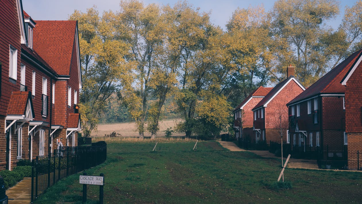 A quiet residential street lined with red-brick houses, bordered by a grassy area and tall trees with autumn foliage under a clear sky. A sign reading "Cascade Way" is visible in the foreground.