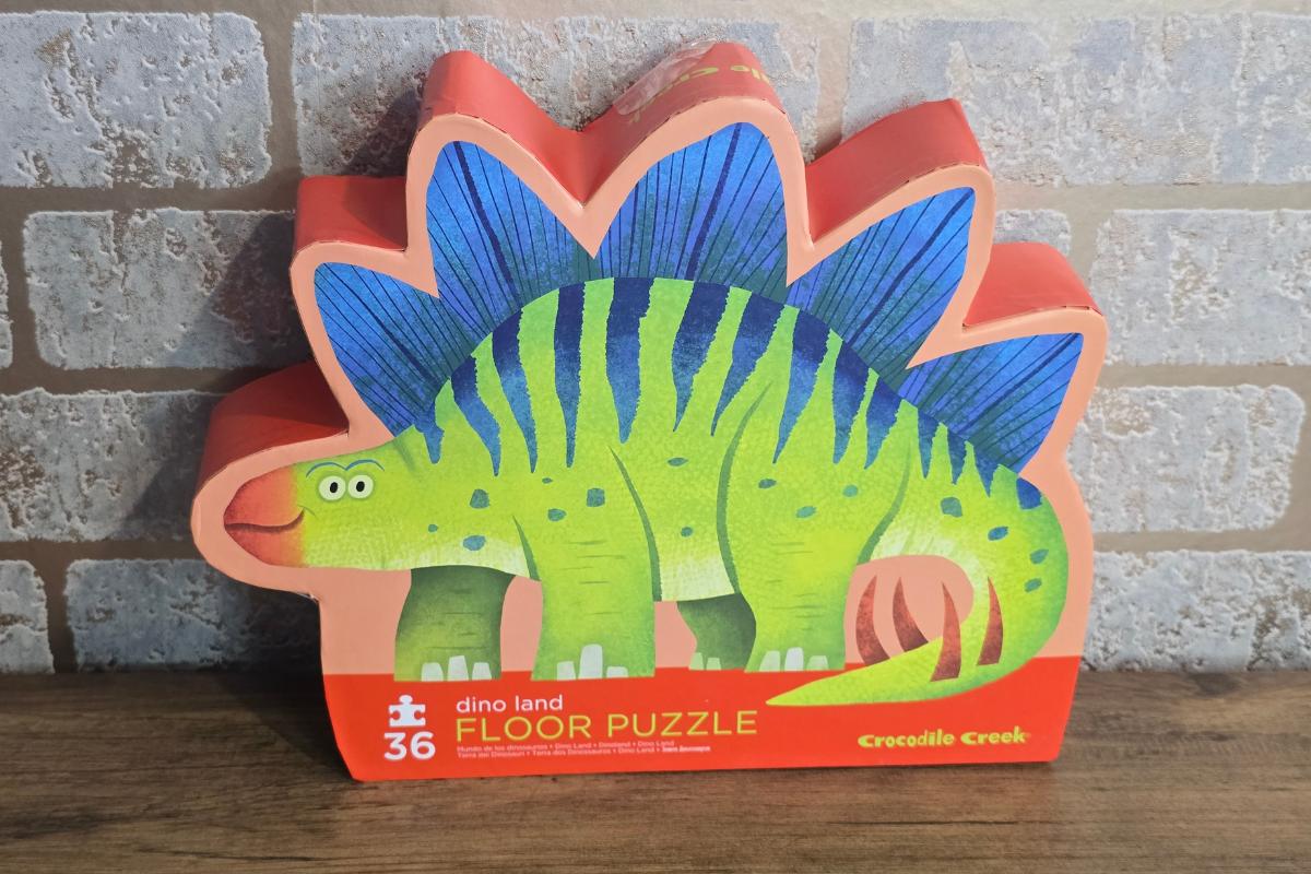 A colorful box shaped like a stegosaurus contains a 36-piece "dino land floor puzzle" by Crocodile Creek—an imaginative pick for children's Christmas gifts, featuring a green dinosaur with blue and orange plates against a brick wall background.