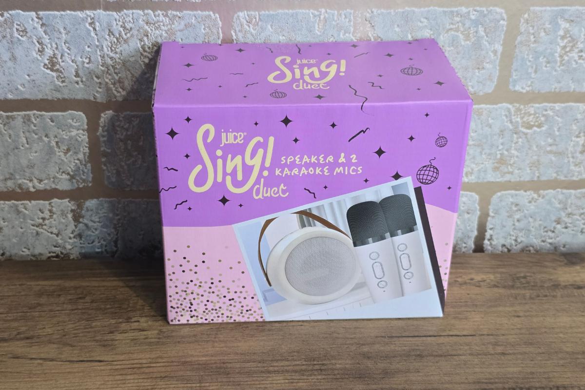 A purple and pink box labeled "Juice Sing! Duet" sits on a wooden surface, perfect for any Children's Christmas gift guide. It features images of a white speaker and two black karaoke microphones, with a brick wall in the background.