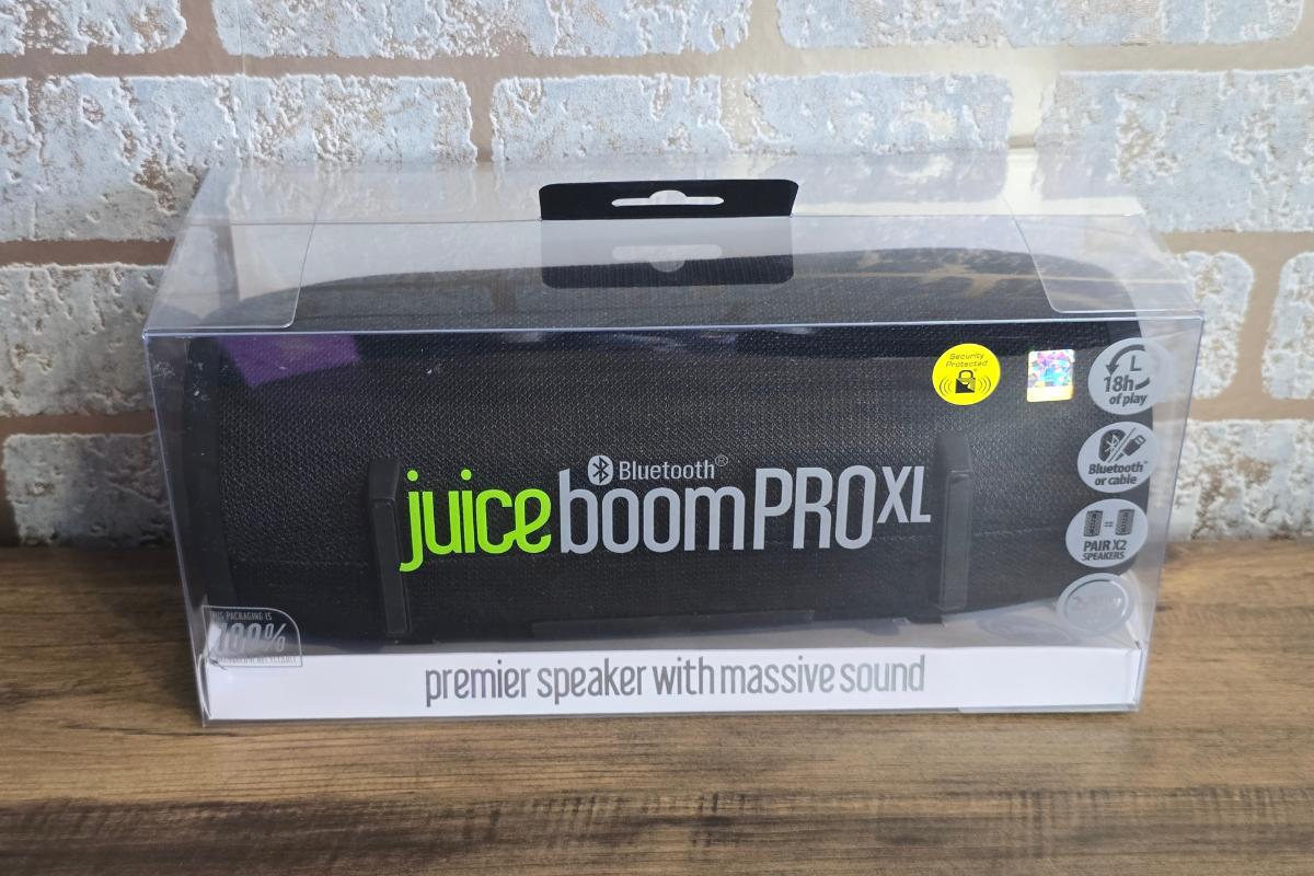 A Juice Boom PRO XL Bluetooth speaker in black packaging sits on a wooden surface against a brick wall background. Perfect for Christmas Gifts, the box highlights features like 18-hour playtime, Bluetooth, and powerful sound.
