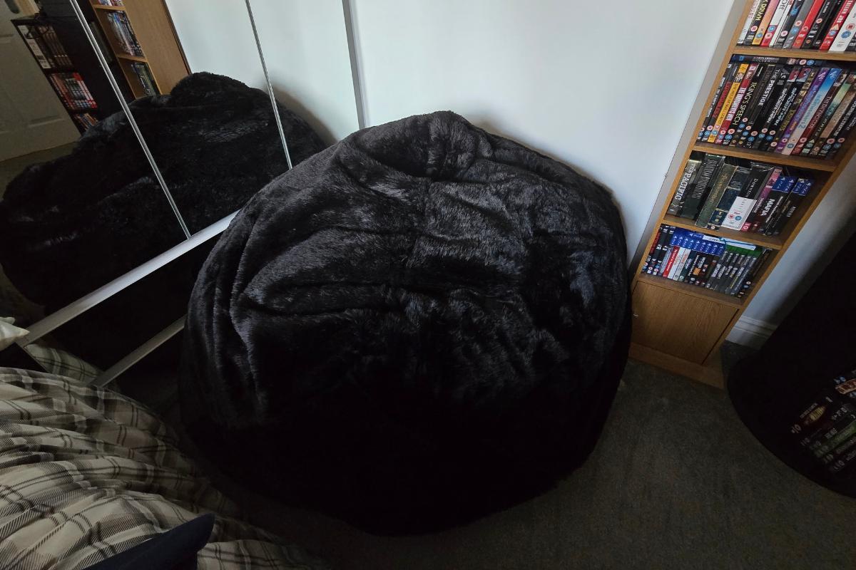 A large black Monster Bean Bag from GreatBeanBags sits in the corner of a room next to mirrored wardrobe doors, a bed with plaid bedding, and bookshelves filled with DVDs and books.