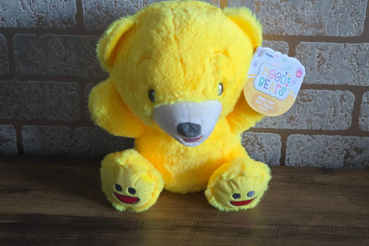 A bright yellow plush teddy bear with a white snout sits on a wooden surface in front of a brick wall. Perfect for children's Christmas presents, this bear has a "Mood Bears" tag and smiling faces stitched on its feet.