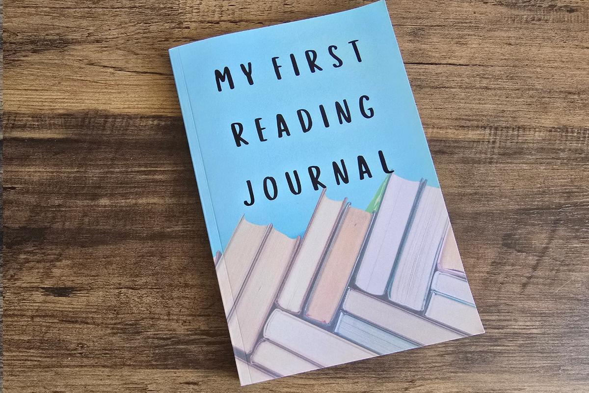 A blue book titled "My First Reading Journal" with a cover illustration of stacked books at the bottom, placed on a wooden surface—perfect for children's gifts or adding to your Christmas gift guide.