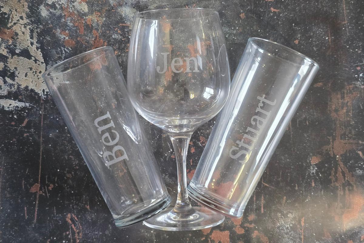Three engraved glasses on a textured, dark surface: a wine glass labeled "Jen" stands in the center, flanked by two tall glasses for "Ben" and "Stuart"—perfect picks from the Ultimate Gift Guide or as thoughtful Gifts for Him.