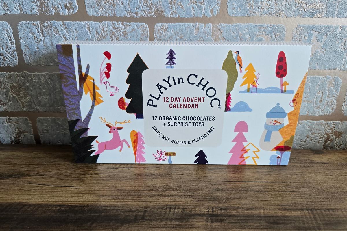 A colorful box labeled "PLAYinCHOC 12 Day Advent Calendar" featuring illustrated trees, a deer, and a snowman. With 12 organic chocolates and surprise toys, it's perfect for children's Christmas gifts—dairy, nut, gluten, and plastic free.
