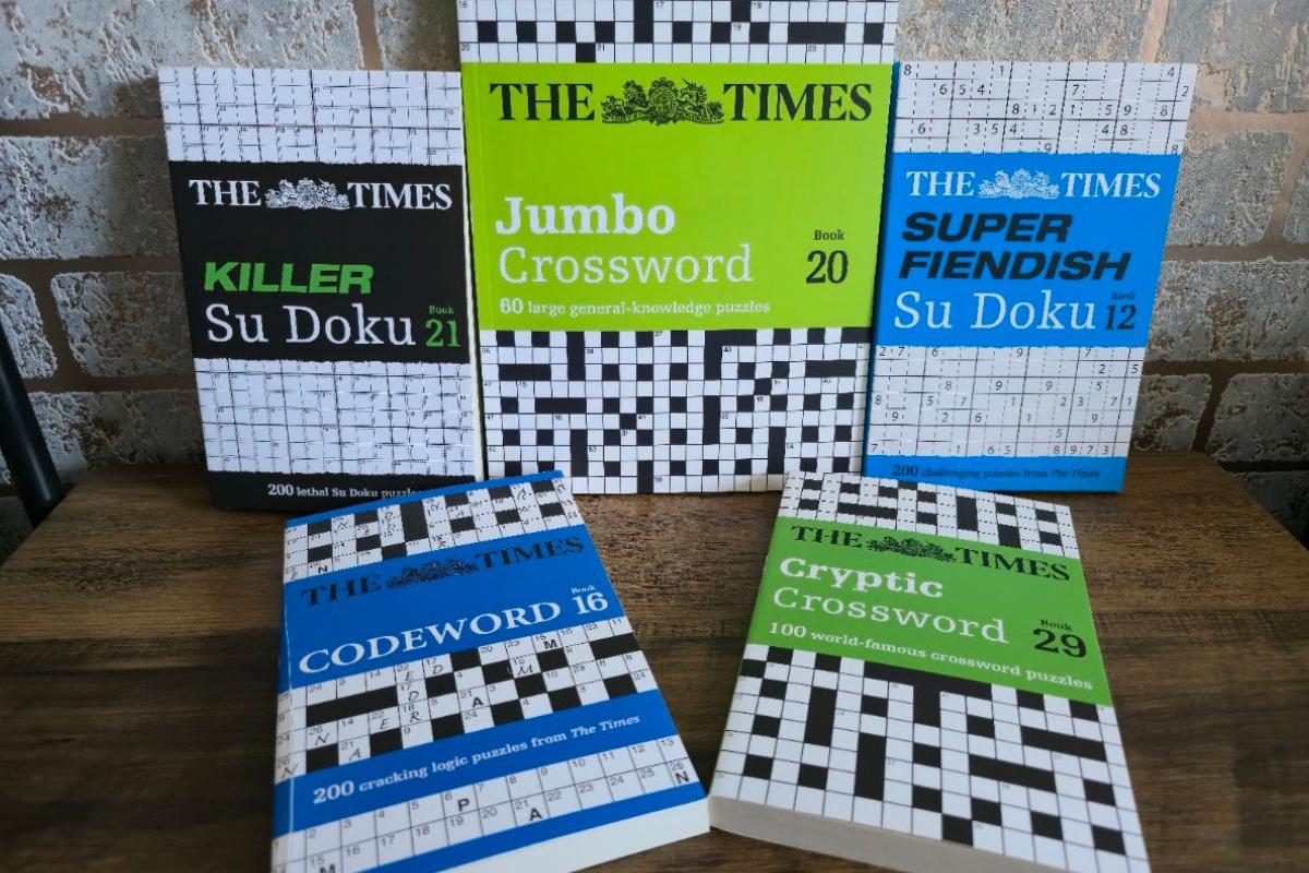 Five puzzle books, perfect for any Gift Guide or Stocking Filler, are arranged on a wooden surface. Titles include Killer Su Doku, Codeword, Jumbo Crossword, Cryptic Crossword, and Super Fiendish Su Doku from "The Times," with a tiled wall behind.